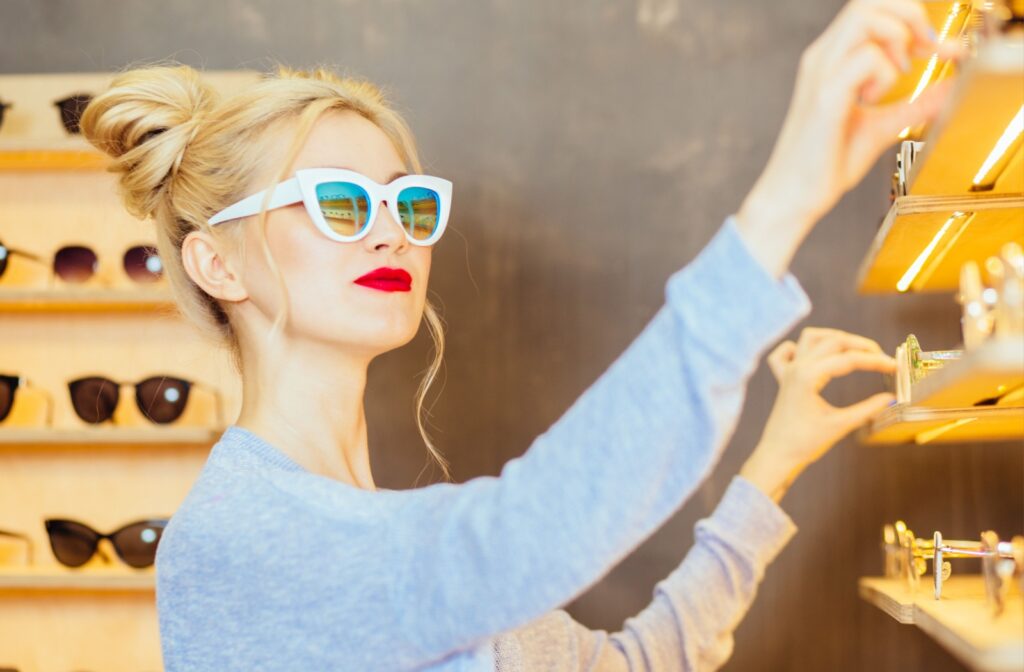 Happy blonde fashionable woman in sunglasses with red lipstick choosing eyeglasses in display of optician store. Consumerism concept.