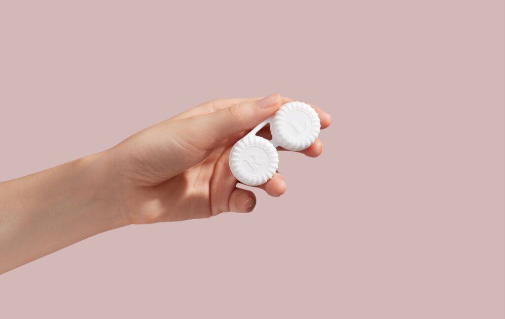 A hand holds out a white contact lens case in the centre of the image in front of a pink background.