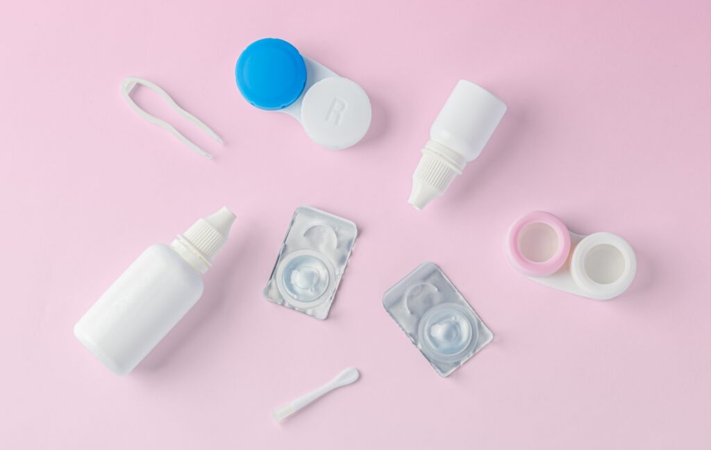 Some daily-disposable contact lenses, a contact lens case, and some bottles of contact lens solutions are arranged across a pink background.