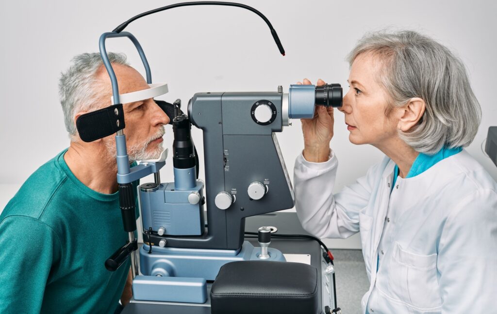An older adult places his head in a slit lamp during a comprehensive eye exam while his optometrist examines his retina.
