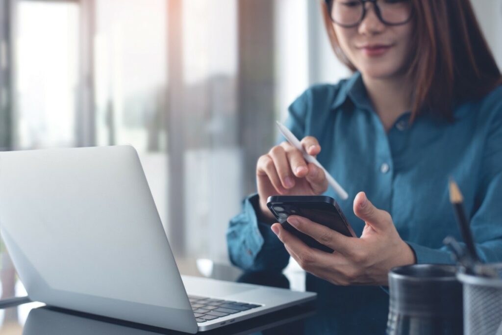 A woman with glasses sitting at a desk, using a white stylus to interact with her smartphone while a laptop is open in front of her.