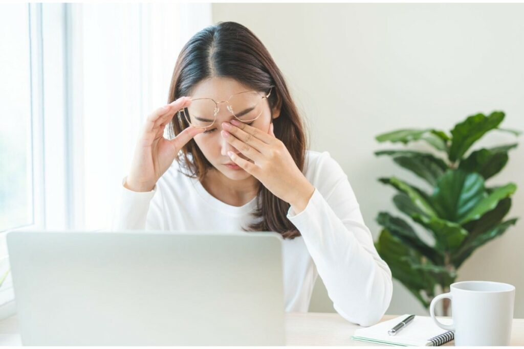 A young Asian woman wearing glasses sitting at a desk with a laptop, looking visibly stressed and tired while rubbing her eyes, suggesting symptoms of digital eye strain or computer vision syndrome.